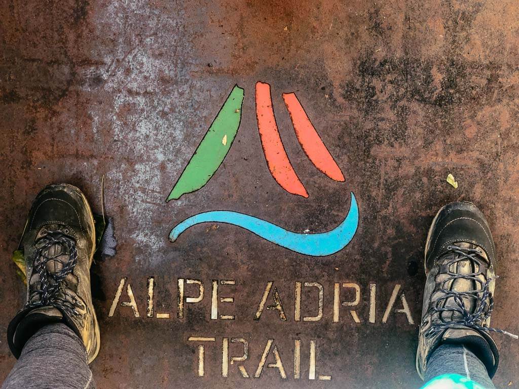 Couchflucht in Wanderschuhen auf dem Schild Alpe-Adria-Trail